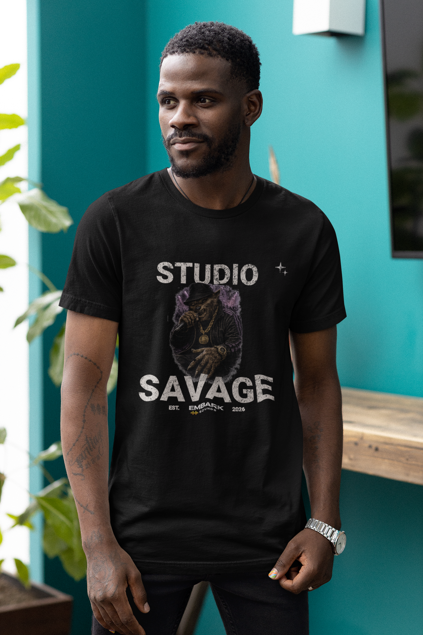 Man wearing a black t-shirt with 'Studio Savage' graphic design against a teal wall.